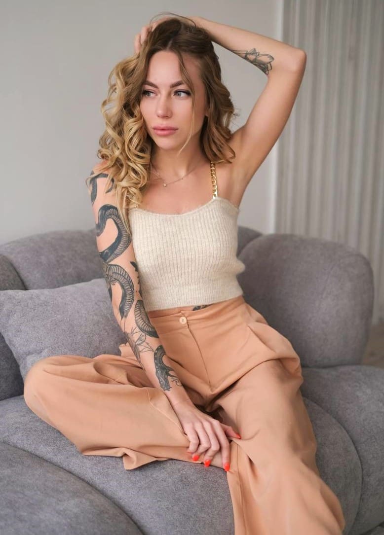 A tattooed woman sits on a gray sofa in a cream ribbed camisole and tan trousers, with one hand raised to her head.