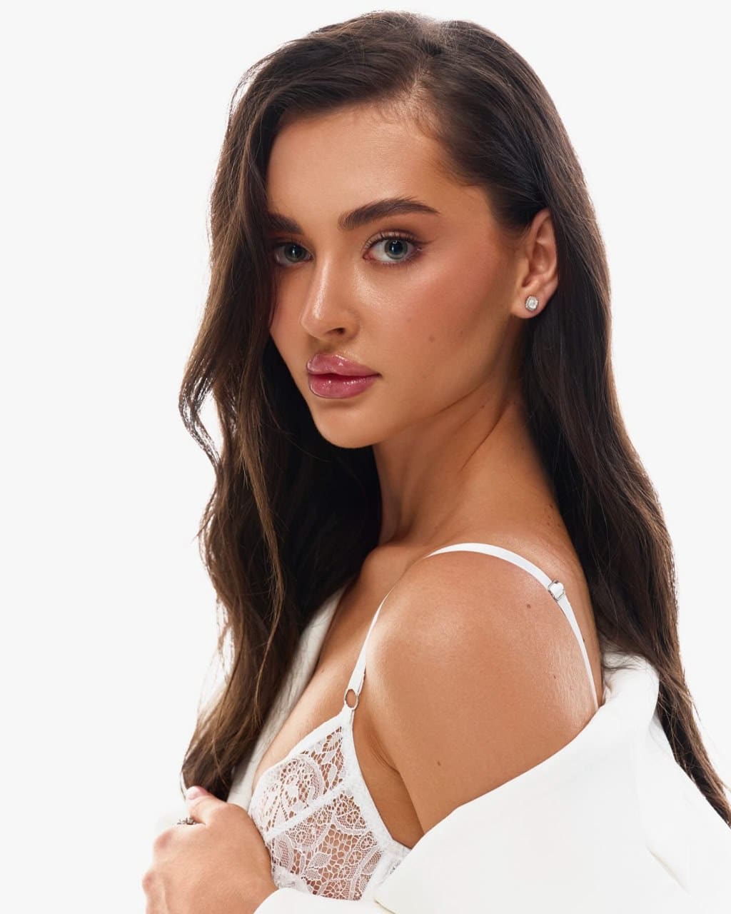 Angelika with long dark wavy hair wears a white lace bralette and off-shoulder jacket against a plain light background.
