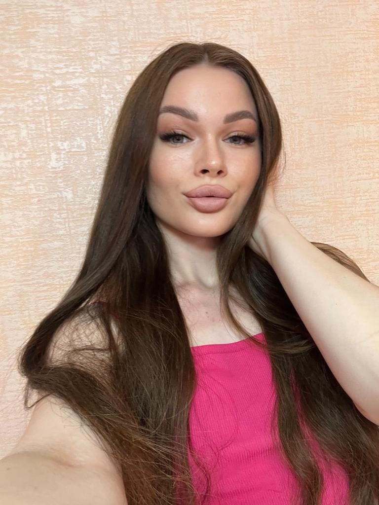 Stone poses in a close-up selfie, wearing a sleeveless pink top with one hand behind her head against a textured beige wall.