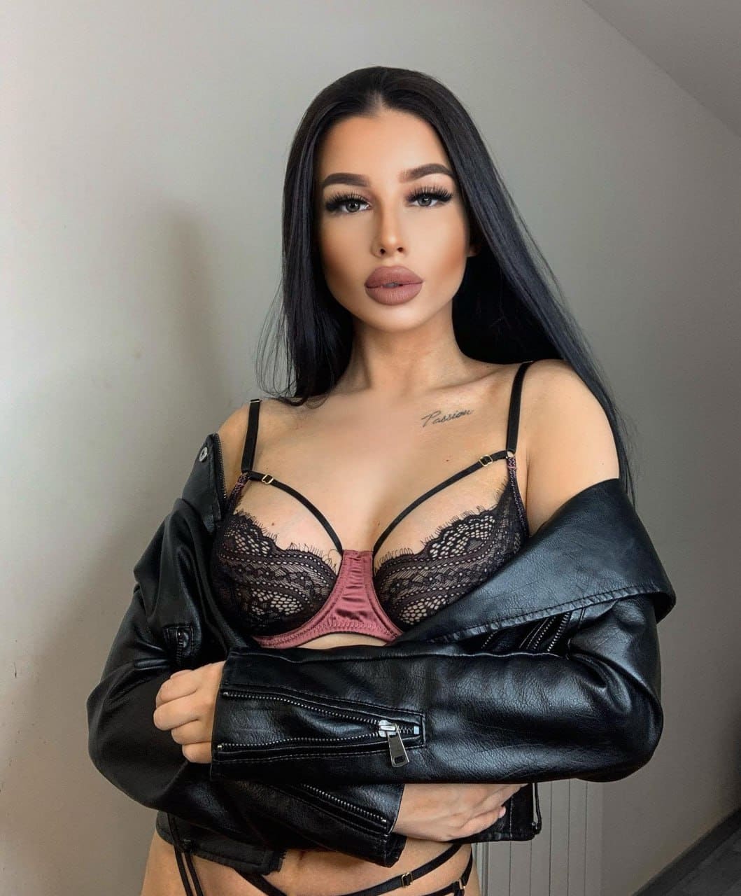 Ashley stands against a plain wall in a black lace bra and off-shoulder leather jacket, arms crossed.
