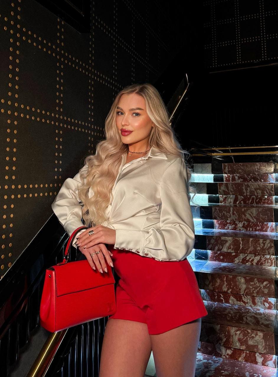 Demidel stands on a marble staircase in a cream satin shirt and red shorts, holding a red handbag.