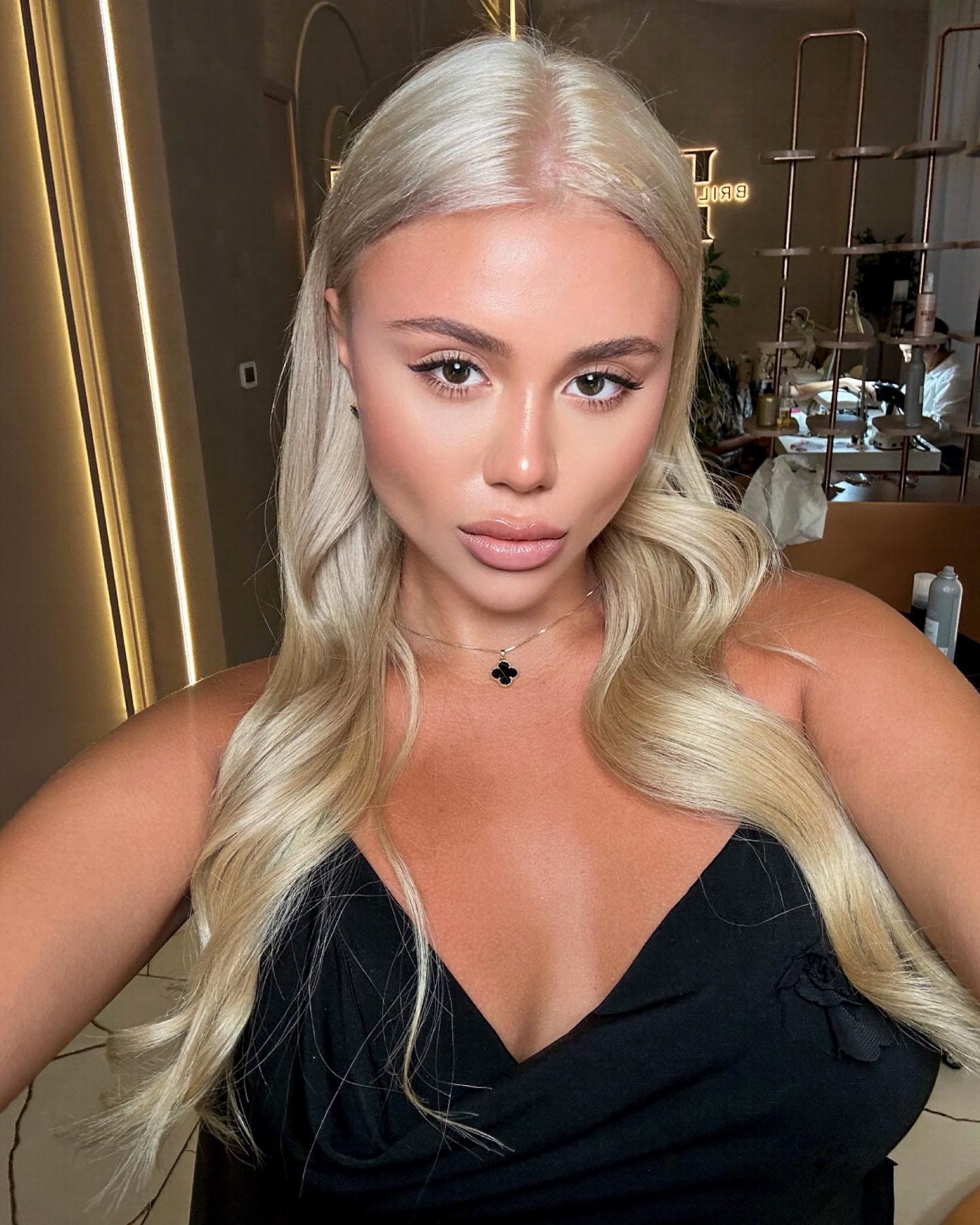 Marta wears a black sleeveless top with a deep V-neck and pendant, posing in a selfie indoors near shelves and a neon sign.