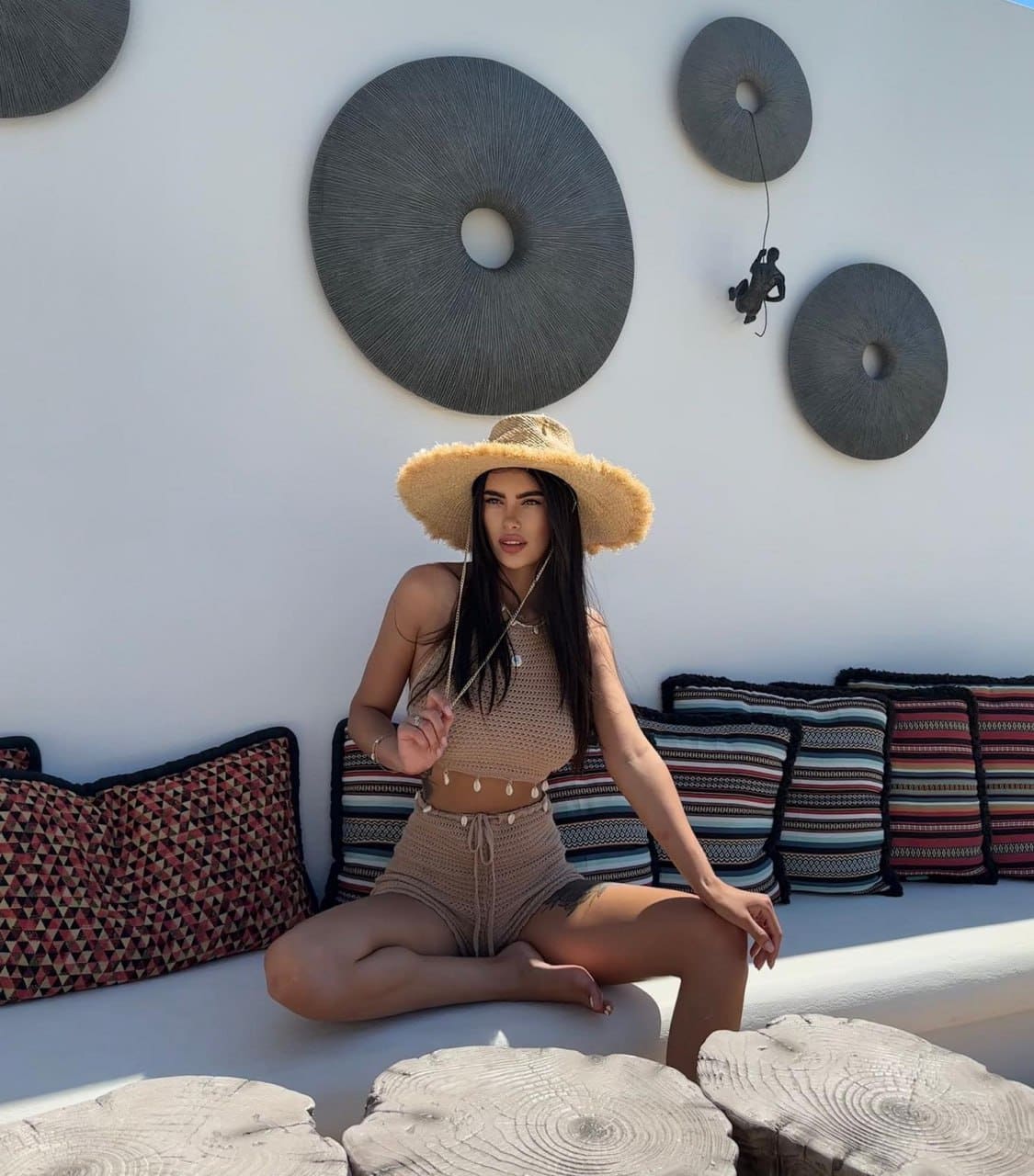 A woman sits cross-legged on a white bench in a tan crochet outfit and straw hat, with patterned cushions behind.