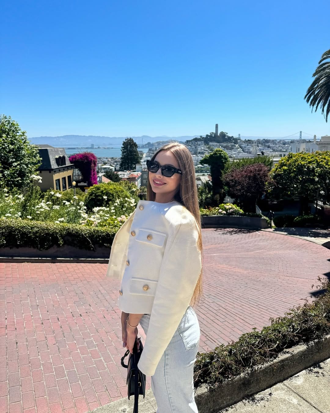 A woman stands on a brick terrace in a white jacket, jeans, and sunglasses, holding a black handbag, with a cityscape and blue sky behind.