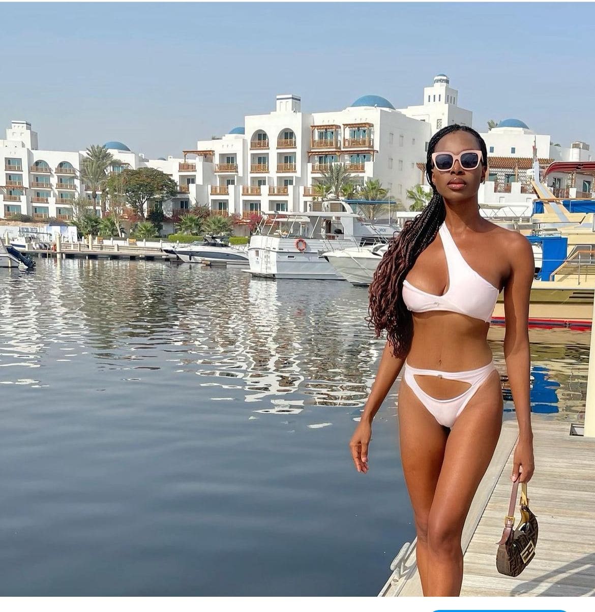 A woman with long braids stands on a wooden dock in a white one-shoulder bikini and sunglasses, holding a handbag near boats.
