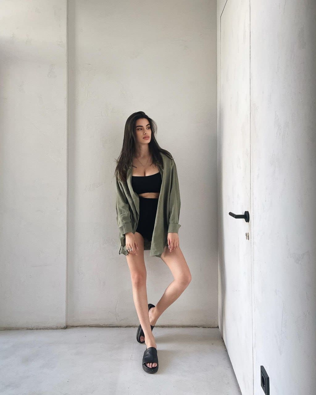 A woman stands in a white room wearing a black bandeau, shorts, and an open olive shirt, with one leg crossed.