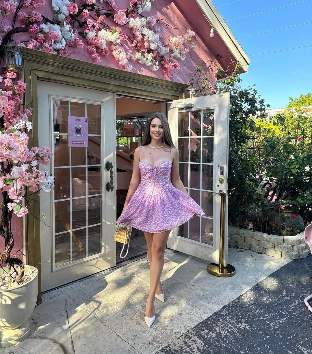 A woman stands before a pink entrance with flowers, wearing a purple floral dress, white heels, and pearl necklace, holding a woven bag.