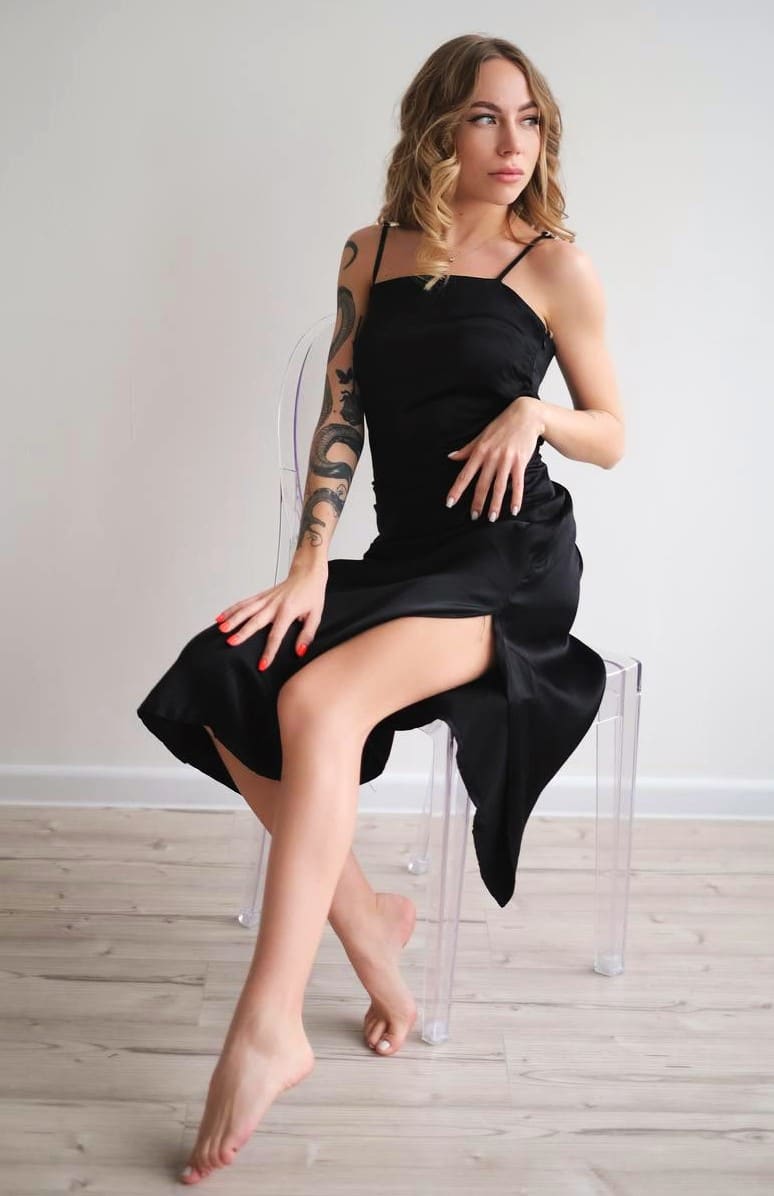 Barefoot on a clear acrylic chair, a woman wears a black satin slip dress with a high slit against a white wall and wood floor.