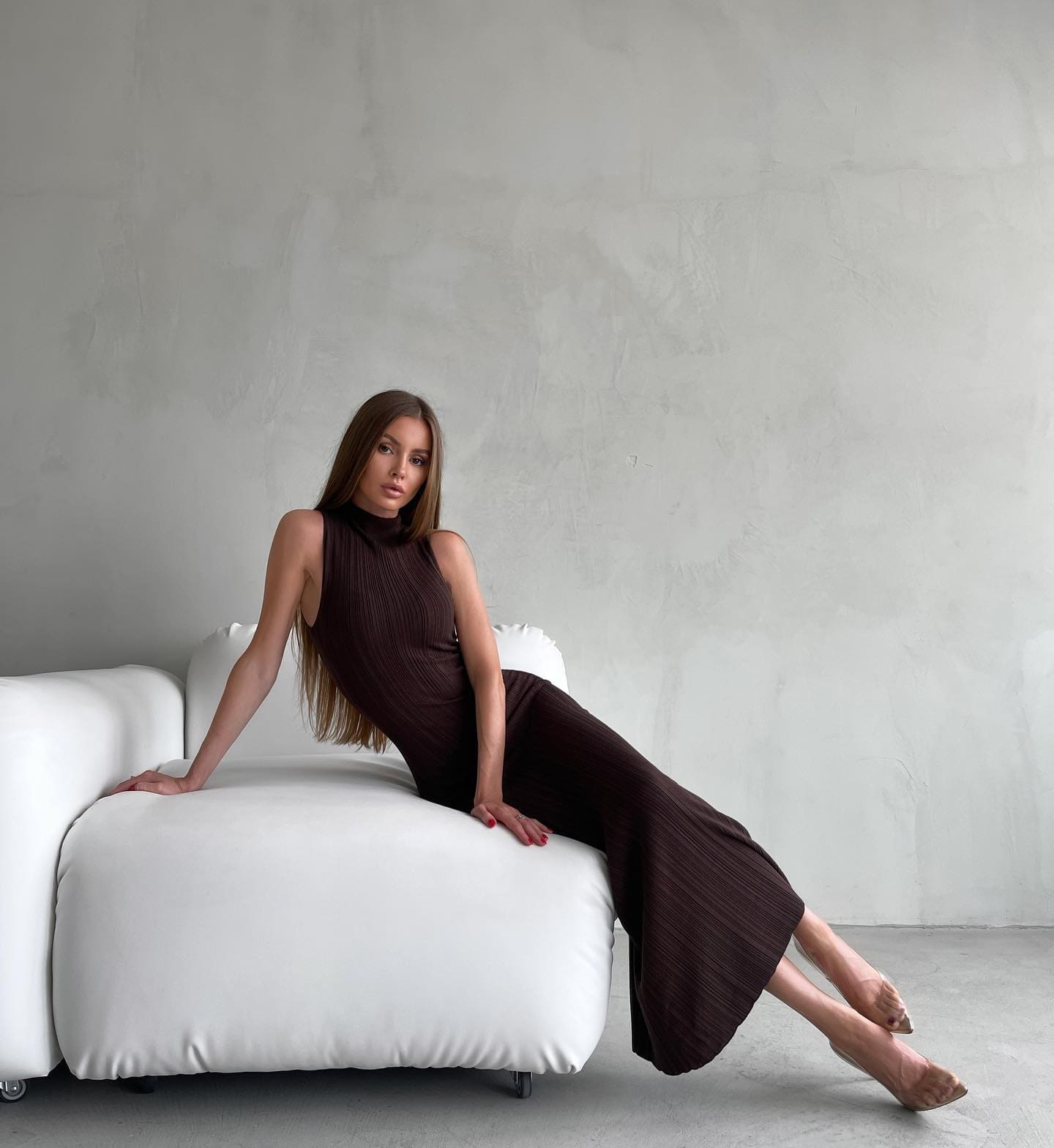A woman sits on a white sofa in a dark brown ribbed dress and clear heels, legs extended to one side against a plain gray wall.