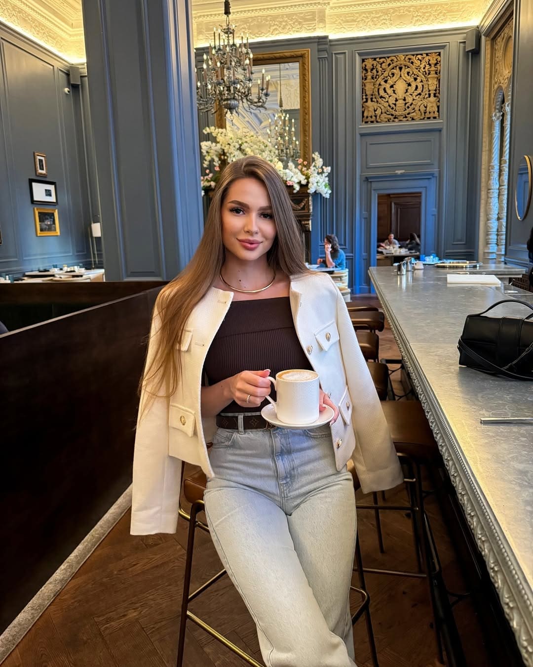 Victoria sits on a stool in an ornate restaurant, wearing a white cropped jacket, dark top, and jeans while holding a coffee cup.