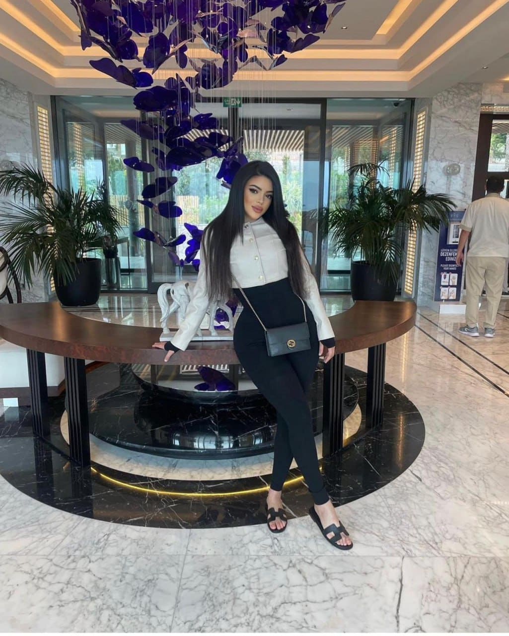 A woman in a white button-up shirt and black leggings poses in a marble lobby beside a wooden table under a chandelier.