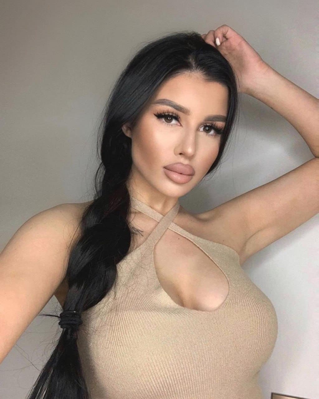 With hair in a side braid, she poses in a beige halter top against a plain wall, arm raised to her head.