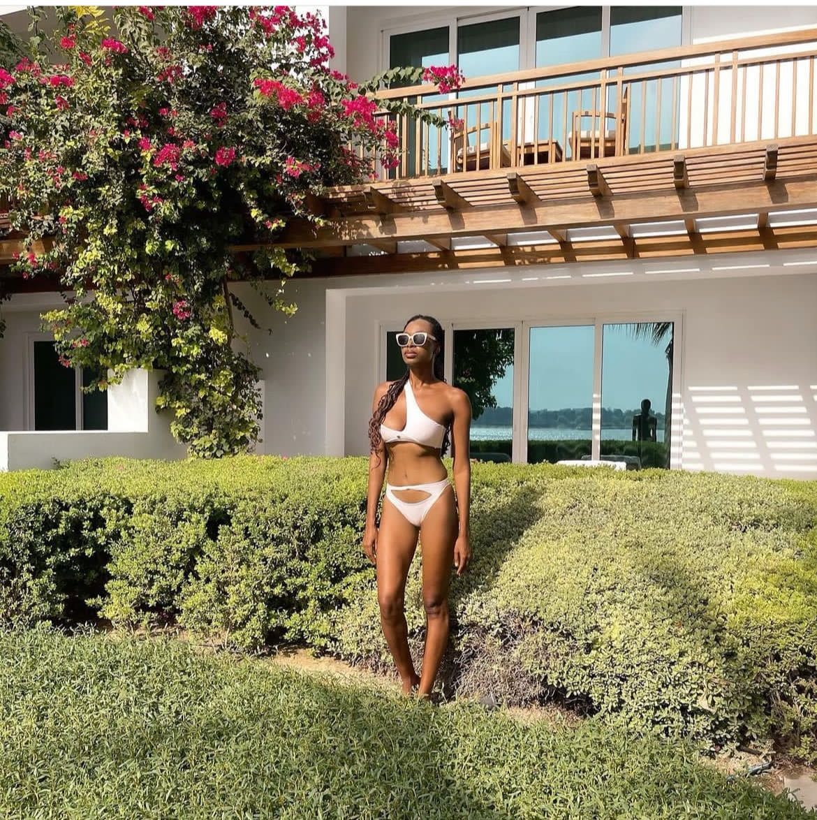 A woman with long dark hair stands in grass before a white building with a balcony, wearing a white cutout swimsuit and sunglasses.