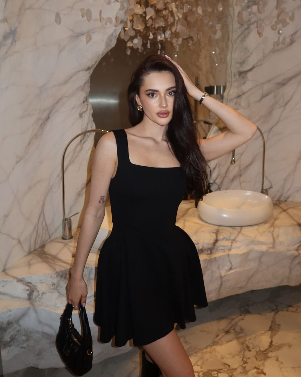 Woman in a black mini dress stands in a marble bathroom, holding a handbag with one hand on her head.