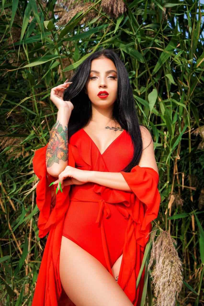Luna stands in red swimwear and an open robe among tall green reeds, one hand touching her long dark hair.