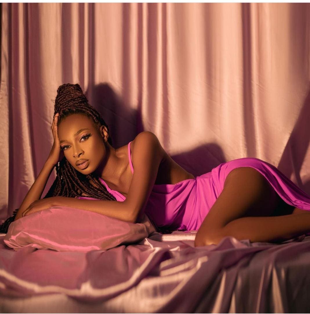 A woman with braided hair in a bun lies on pink satin sheets in a short pink slip dress, hand on head, against pink curtains.