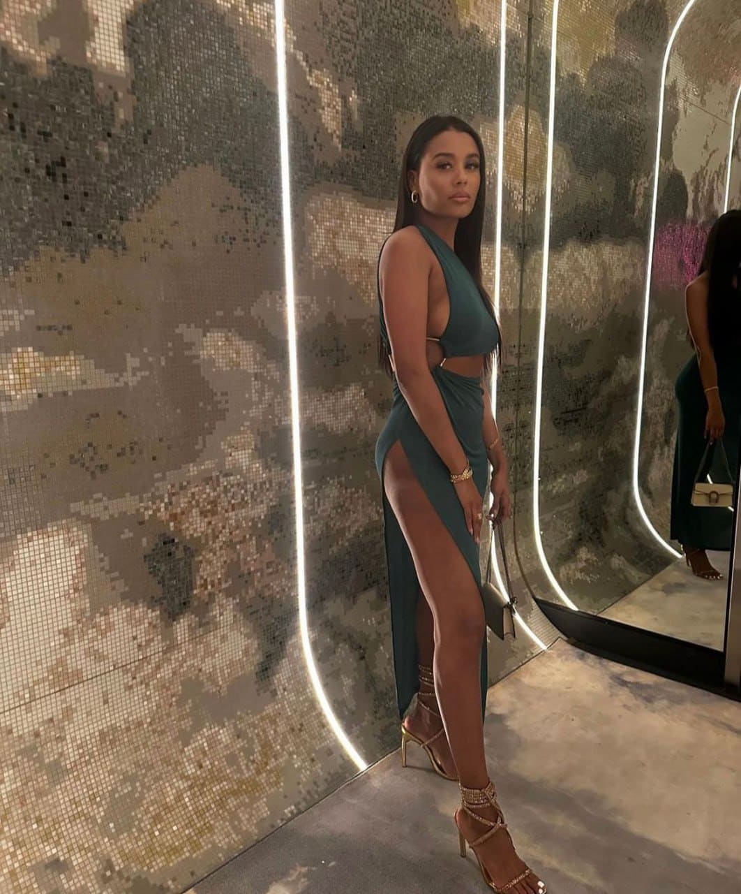 A woman in profile wears a dark green two-piece outfit with a high-slit skirt, posing before a mosaic wall with light strips.