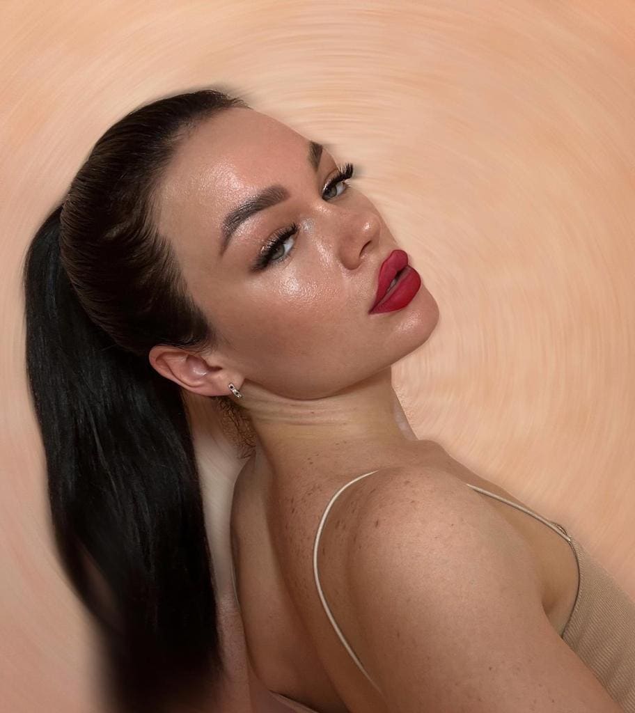 A woman with a high ponytail and red lipstick looks over her bare shoulder, wearing a thin-strapped beige top against a swirled peach background.