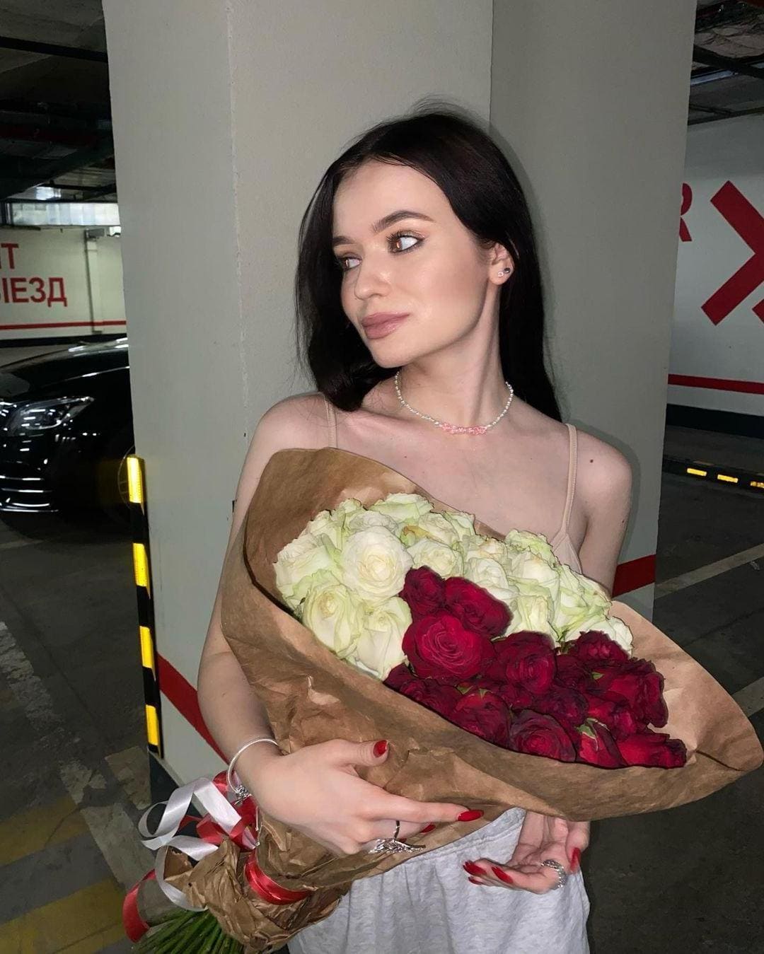 A woman in a light spaghetti-strap top stands in a parking garage holding a bouquet of roses wrapped in brown paper, looking to the side.