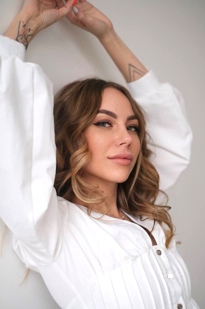 Juli stands with arms raised against a light wall, wearing a white button-up blouse with sleeves pushed up to reveal forearm tattoos.