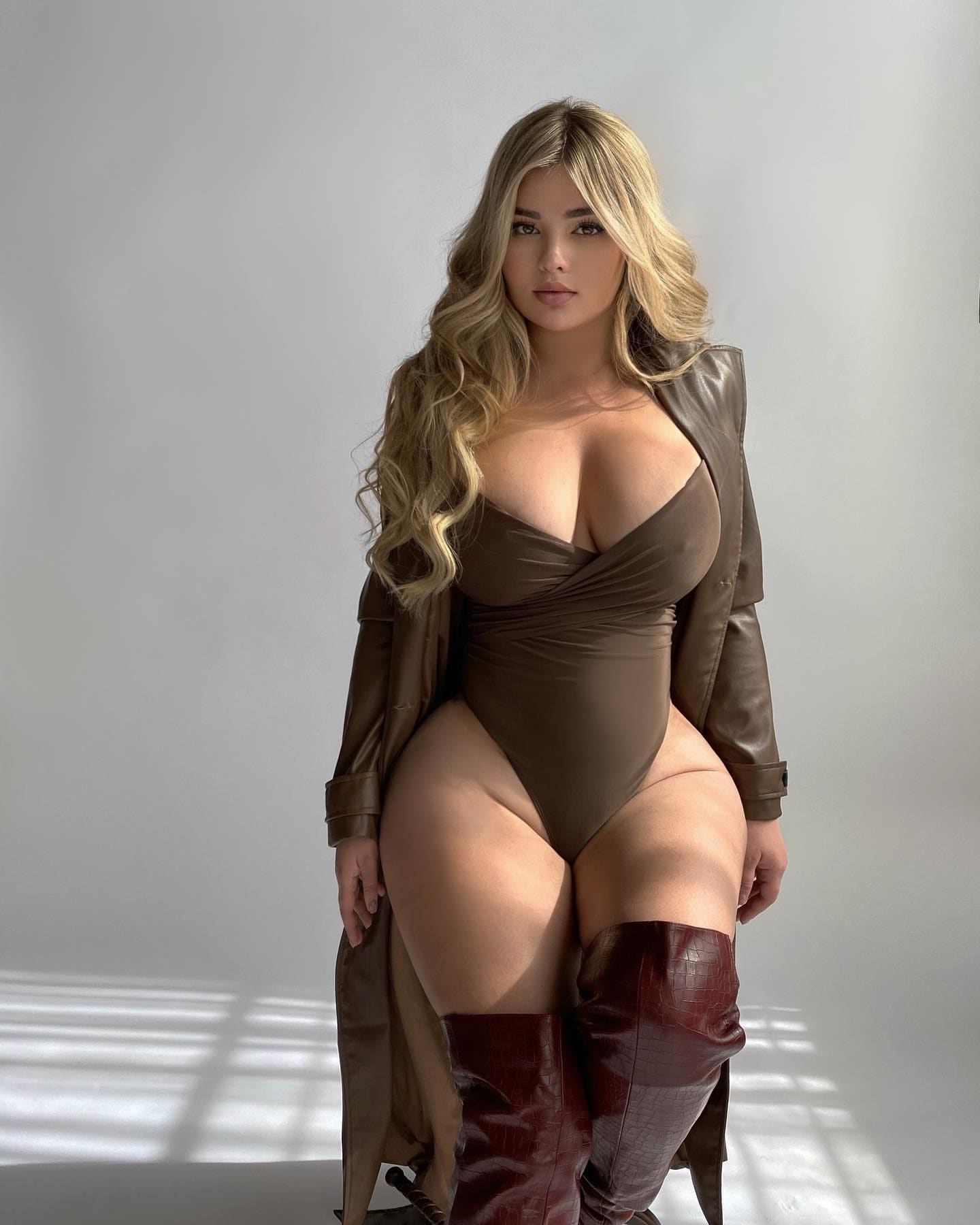 Lilly stands in a studio wearing a brown bodysuit, open leather coat, and knee-high red boots against a plain light background.