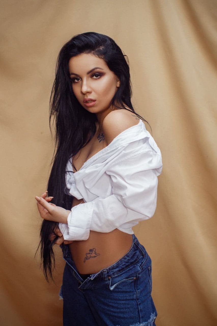 A woman stands against a tan backdrop in an unbuttoned white shirt and low-slung blue jeans, holding her hair.