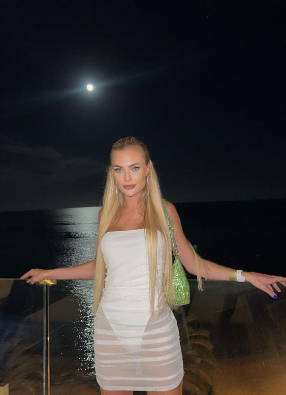 A woman stands at a glass railing at night in a white strapless mini dress with sheer panels, a full moon behind her.