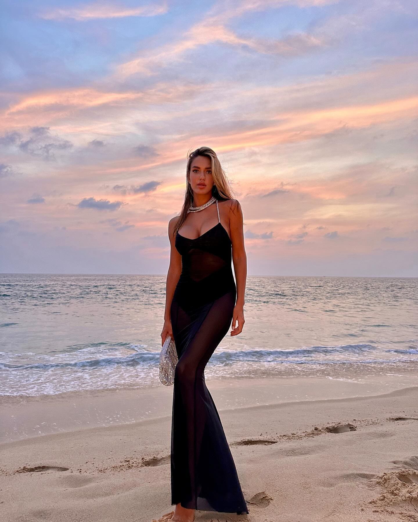 A woman in a long black dress with sheer panels stands on a beach at sunset, holding a silver clutch.