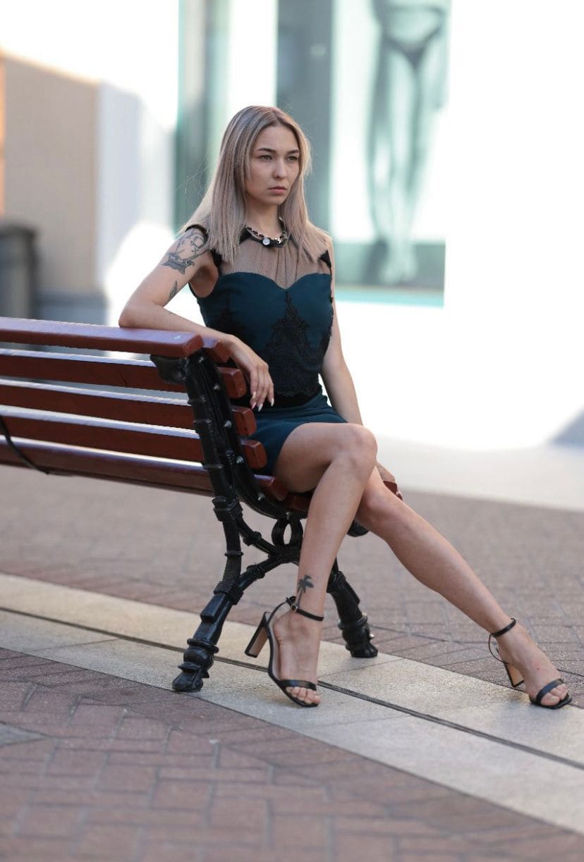 A blonde woman sits on a wooden bench outdoors, wearing a short dark green dress with mesh and lace details, black heels, and visible arm tattoos.