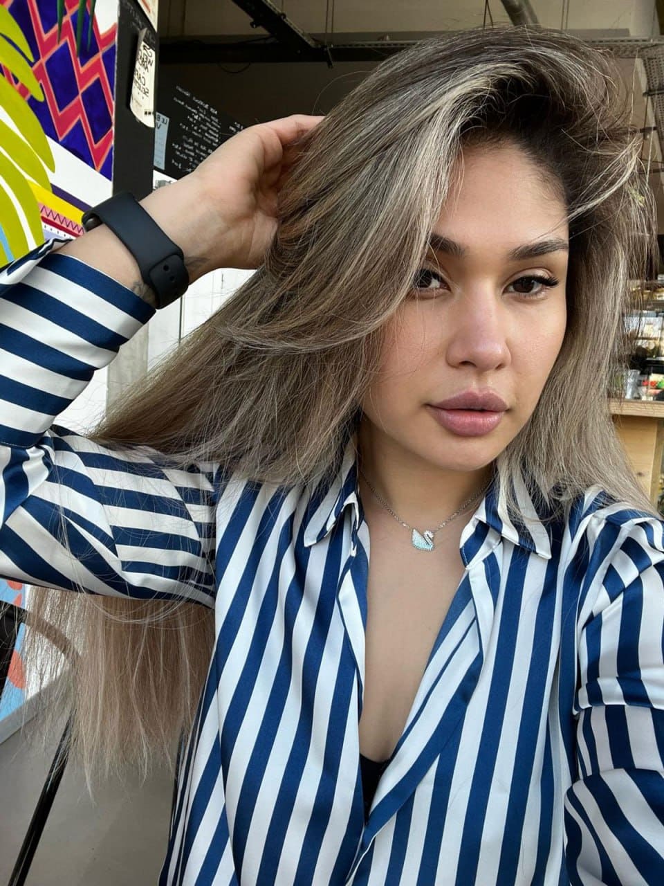 Kristy takes a selfie indoors, wearing a blue and white striped shirt over a black top, with colorful artwork behind her.