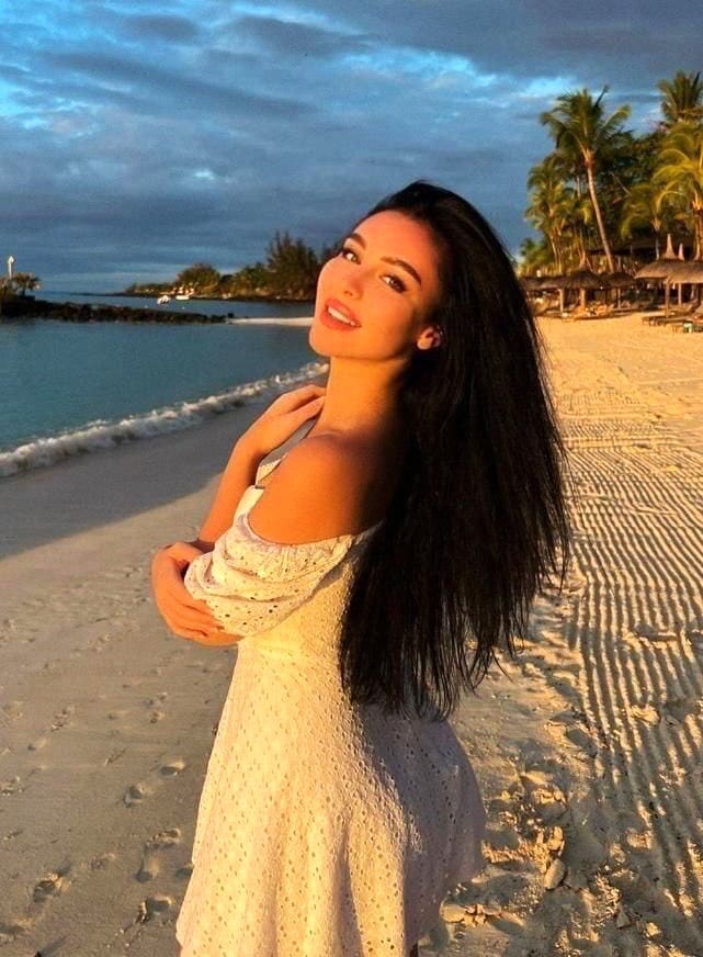 A woman in a white off-the-shoulder dress stands on a sandy beach at dusk, with ocean and palm trees in the background.