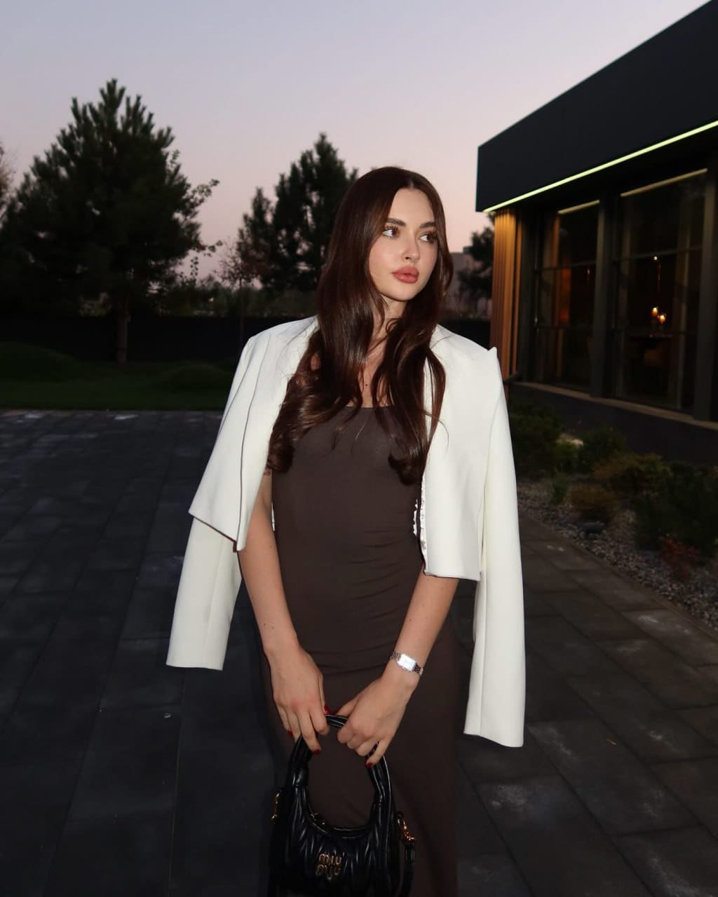 Woman in a brown dress and white cropped jacket stands on pavement at dusk, holding a black handbag.