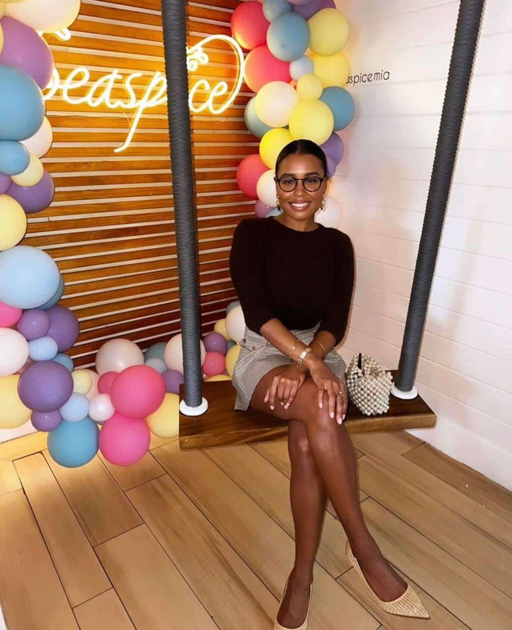 A woman in glasses, a dark top, and plaid skirt sits on a swing bench near a neon sign and balloon arch, with a beaded bag.