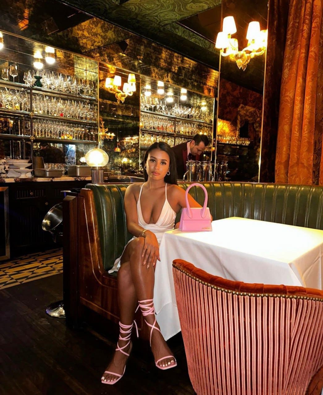 A woman in a white deep-V halter dress and pink heels sits in a green leather booth at a restaurant table with a pink handbag.