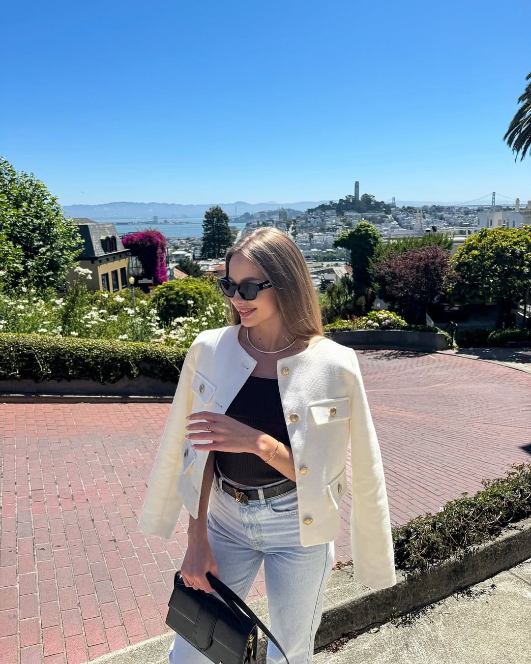 A woman on a brick terrace wears sunglasses, a white cropped jacket, black top, and jeans, holding a black bag against a city and water view.