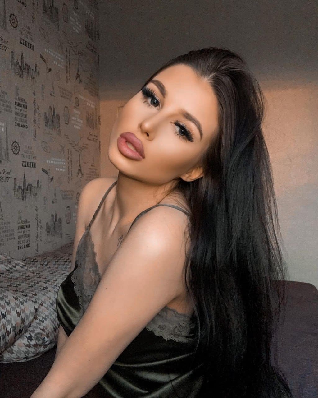 Sitting on a bed in a dark satin camisole, she tilts her head toward the camera against patterned wallpaper.