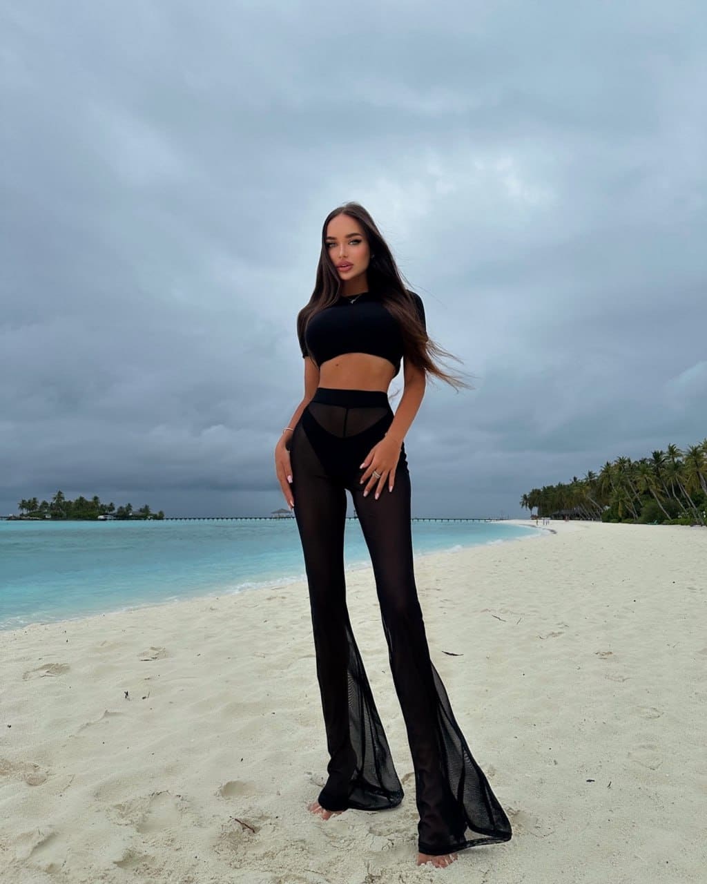 A barefoot woman in a black crop top and sheer flared pants stands on white sand, with turquoise water and palm trees behind her.