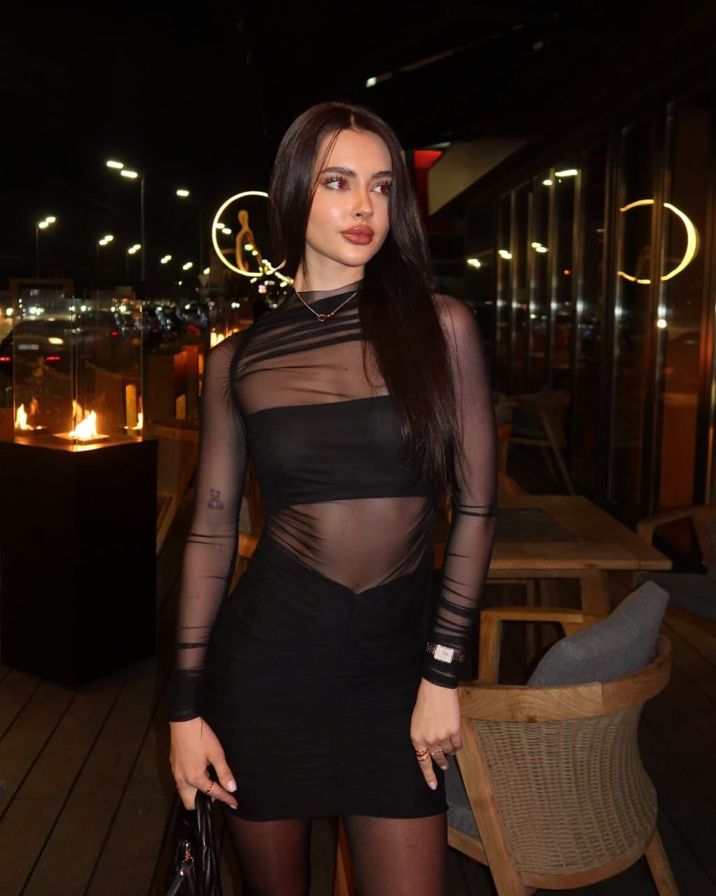 Woman in a black mini dress with sheer mesh sleeves stands outdoors at evening, holding a small bag.