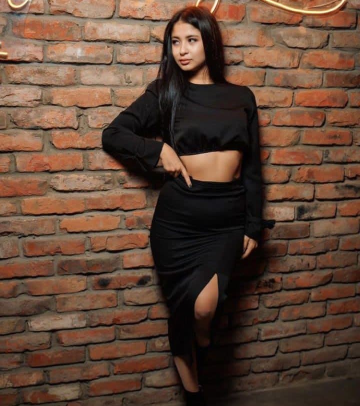 A woman in a black crop top and high-slit midi skirt stands against a red brick wall, one hand resting on her hip.