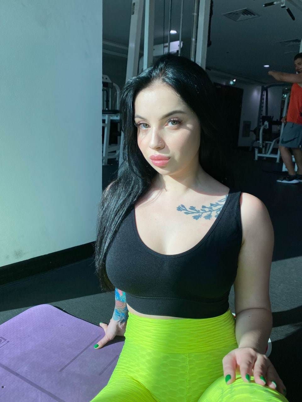 A woman sits on a purple gym mat wearing a black sports bra and green leggings, with visible chest and arm tattoos.