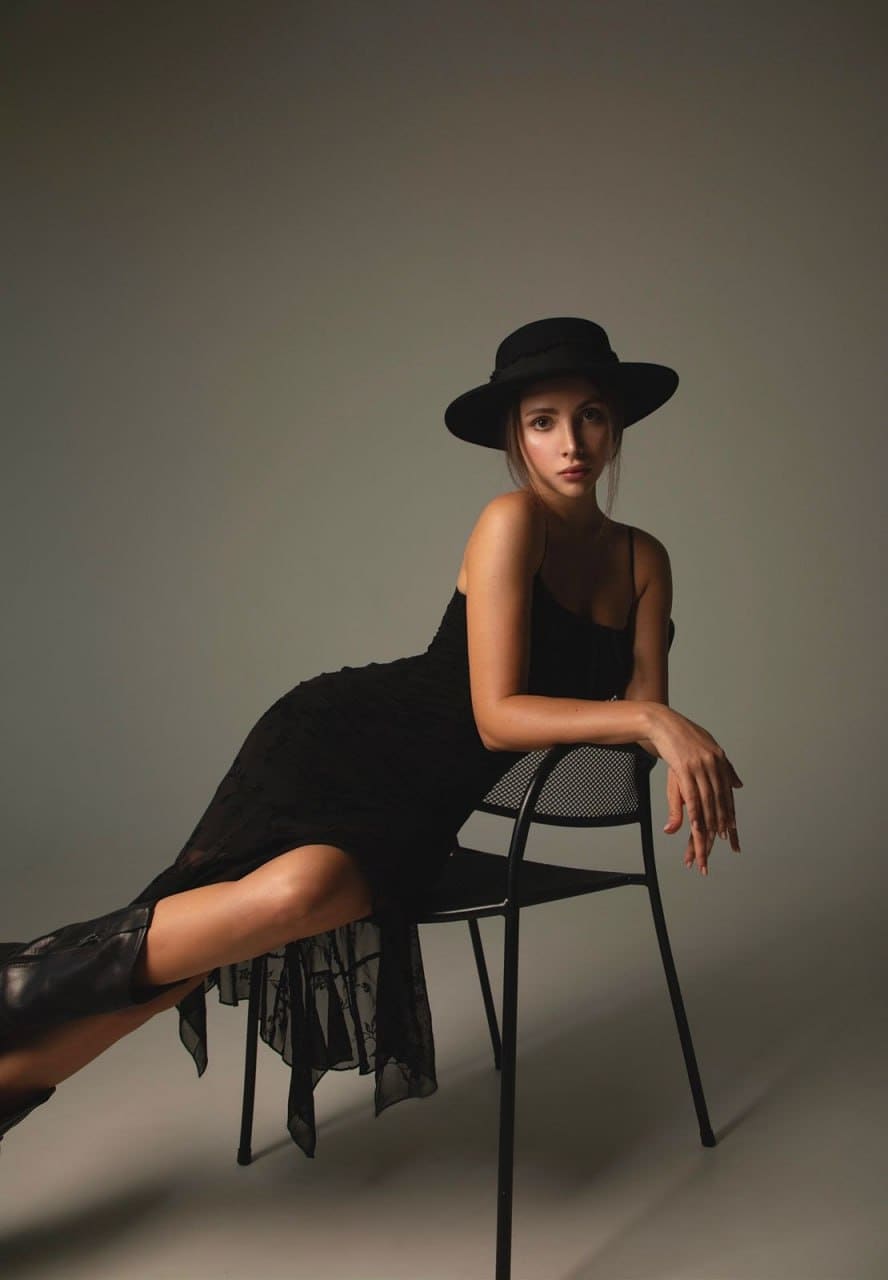 A woman sits on a chair in a black lace dress, hat, and boots against a gray backdrop.