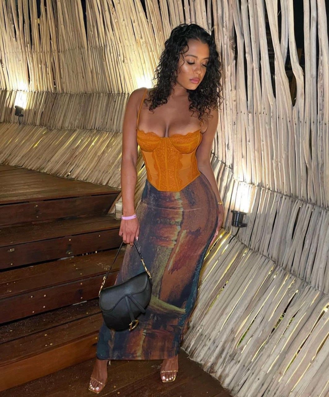 A woman in an orange lace bodice and patterned skirt stands on wooden steps beside slatted walls, holding a black handbag.
