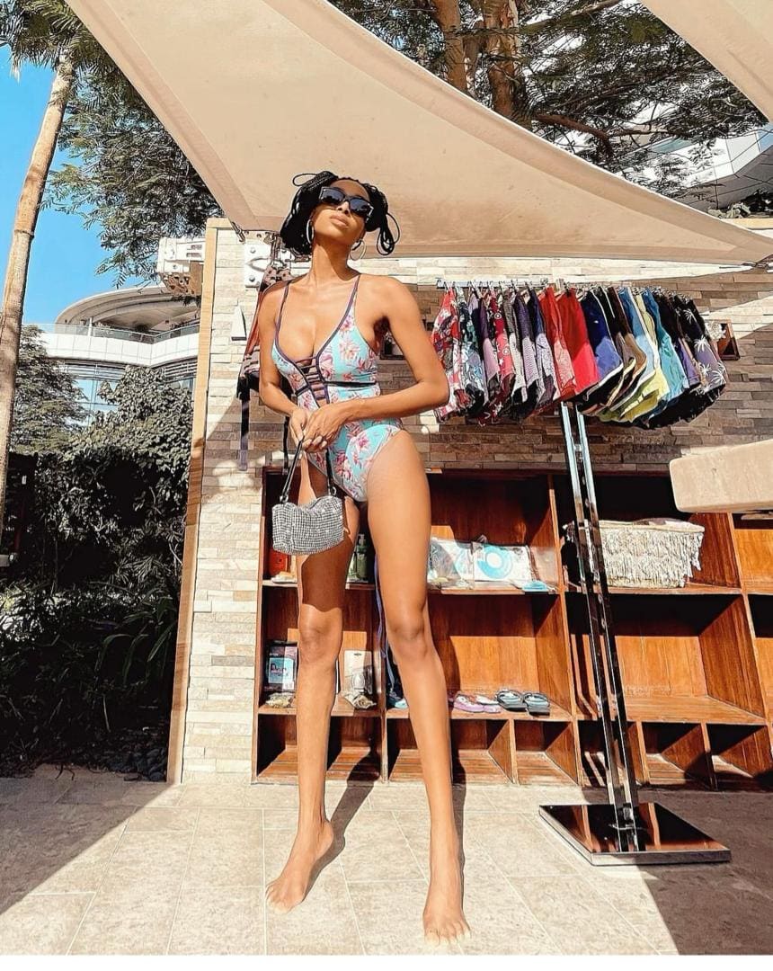 A woman with hair in a bun stands barefoot in a floral one-piece swimsuit and sunglasses, holding a handbag by an outdoor clothing rack.