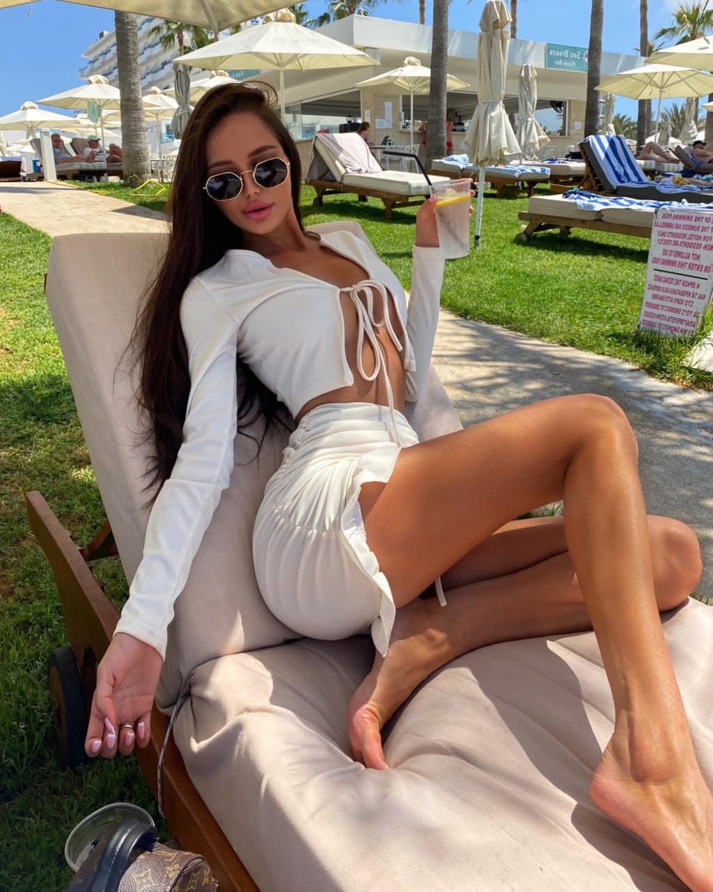 A woman in a white lace-up crop top and mini skirt reclines on a beige lounge chair at a sunny resort, holding a drink.