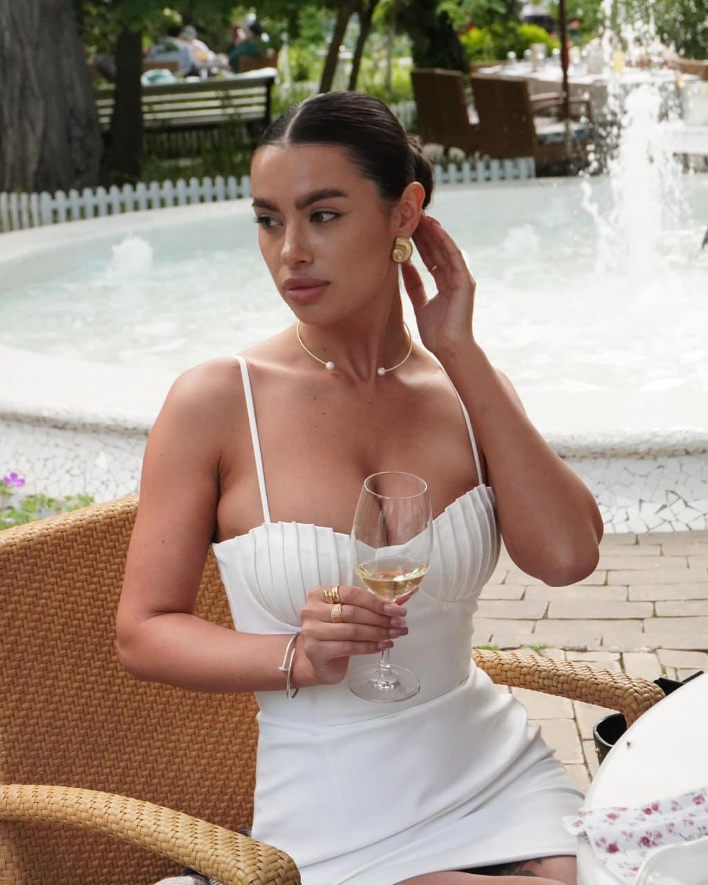 Emily sits in a woven chair outdoors in a white dress, holding a wine glass, with a fountain and greenery behind her.