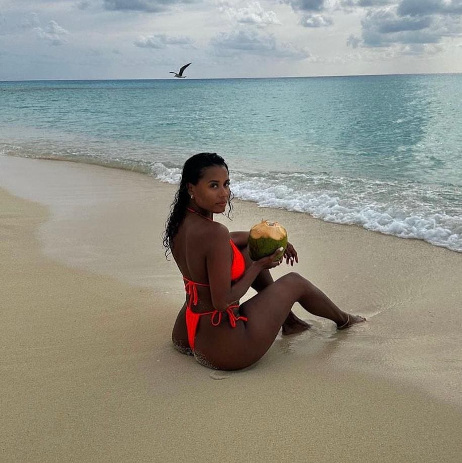 A woman in a red bikini sits on a sandy beach at the water's edge, holding a green coconut and looking back over her shoulder.