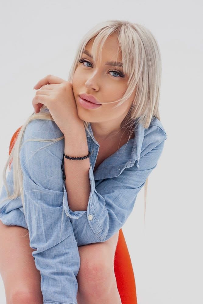 Dari sits on an orange chair in an unbuttoned light-blue denim shirt, hand near her chin, against a white background.