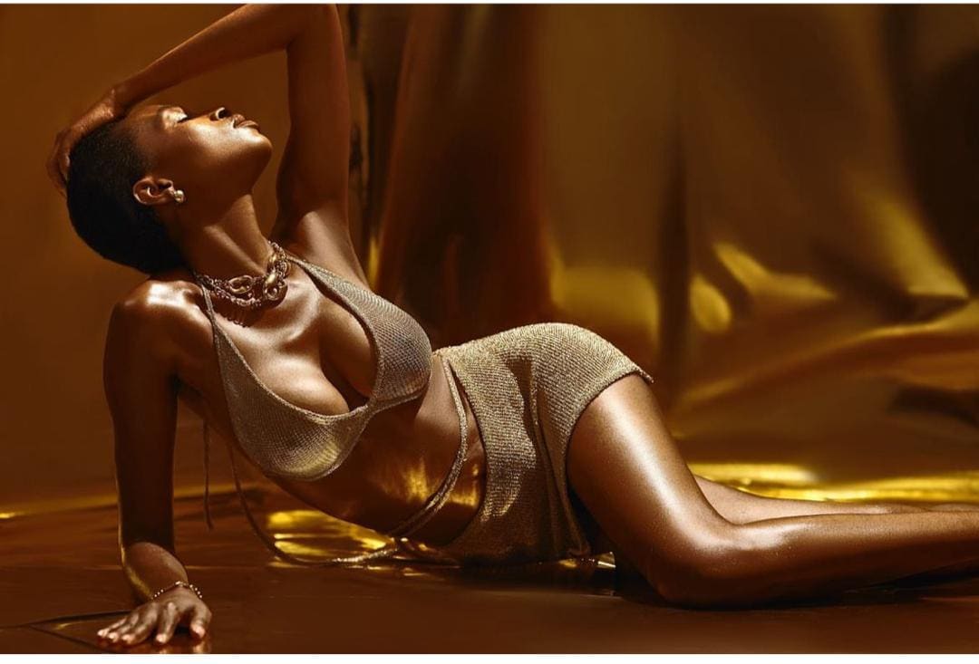A person with short dark hair reclines on a reflective floor in a gold metallic two-piece outfit, arm raised against a golden backdrop.