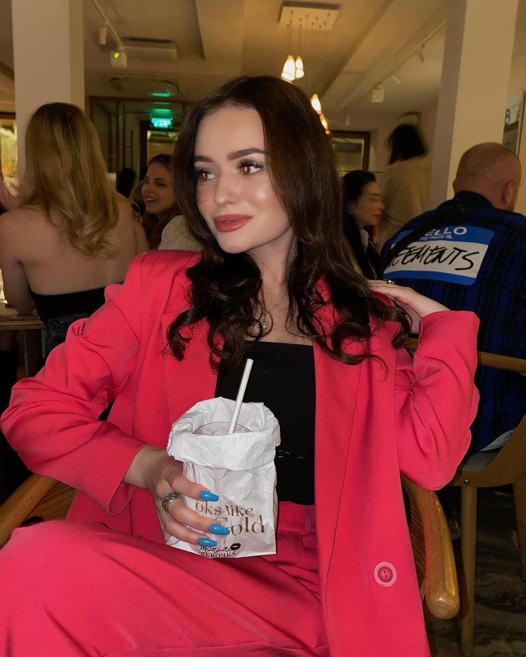 A woman in a bright pink blazer sits in a restaurant holding a drink cup with a straw, looking away from the camera.