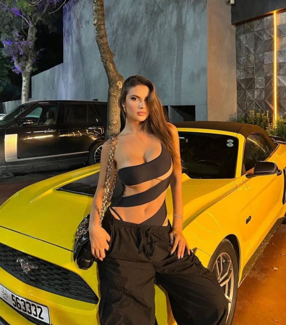 A woman leans against a yellow convertible at night, wearing a black bandeau top, loose trousers, and a chain-strap bag, with an SUV behind.